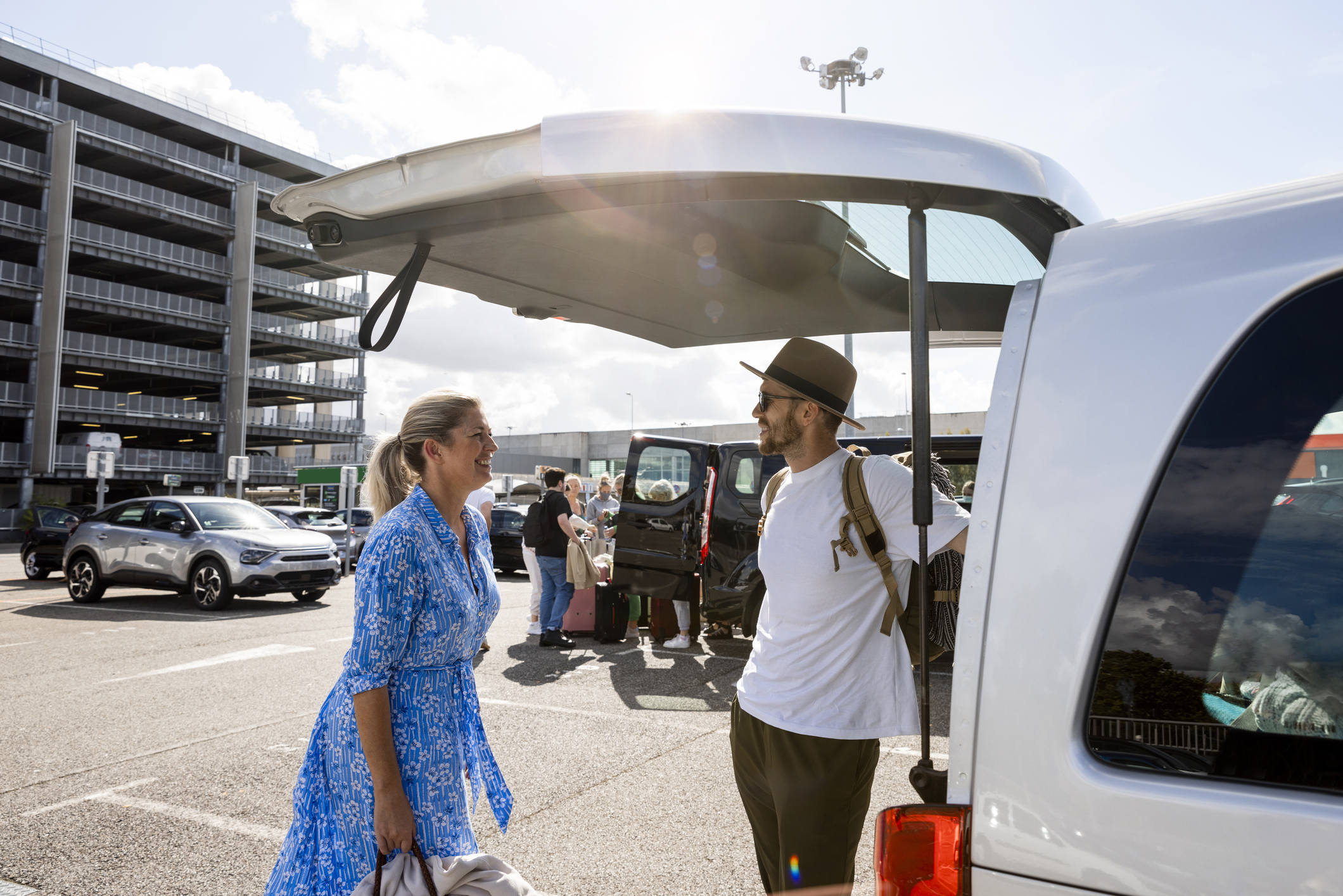 Mother and son packing up a rental van where they are vacationing to. They are standing in a car park at an airport in the sun.
